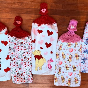 May include: Five hanging kitchen towels with crochet tops. The towels feature Valentine's Day and cartoon character designs, including Snoopy, Winnie the Pooh, and Stitch. The towels have red and pink hearts, and the crochet tops are red or pink.