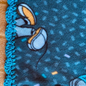 K&ouml;nnte beinhalten: Eine blaue Fleece-Decke mit einem geh&auml;kelten t&uuml;rkisfarbenen Rand. Die Decke zeigt eine Comicfigur mit einem gelben und blauen Design.
