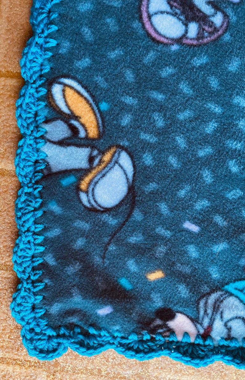 K&ouml;nnte beinhalten: Eine blaue Fleece-Decke mit einem geh&auml;kelten t&uuml;rkisfarbenen Rand. Die Decke zeigt eine Comicfigur mit einem gelben und blauen Design.
