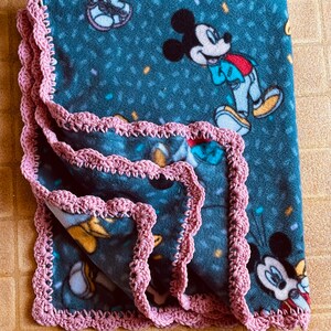 K&ouml;nnte beinhalten: Eine blaue Fleece-Decke mit einem rosa H&auml;kelrand. Die Decke zeigt ein sich wiederholendes Muster von Mickey-Maus-Figuren und Konfetti.