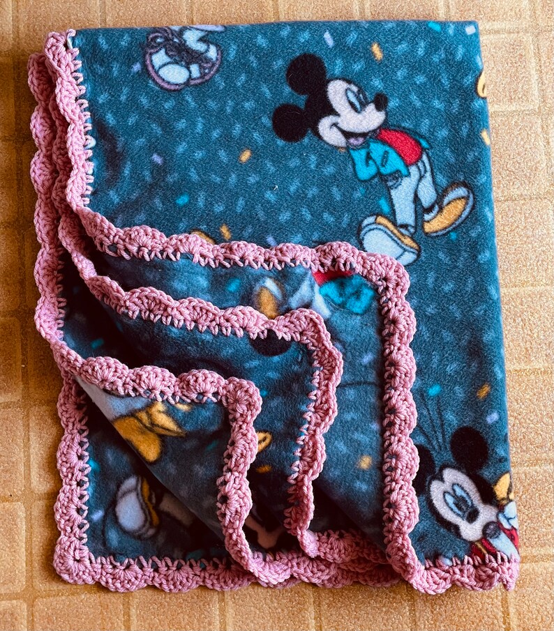 K&ouml;nnte beinhalten: Eine blaue Fleece-Decke mit einem rosa H&auml;kelrand. Die Decke zeigt ein sich wiederholendes Muster von Mickey-Maus-Figuren und Konfetti.