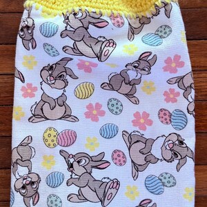 Puede incluir: Un porta-toallas de cocina de crochet amarillo con un bot&oacute;n y una toalla de cocina blanca con un patr&oacute;n de conejos marrones y huevos de Pascua coloridos.