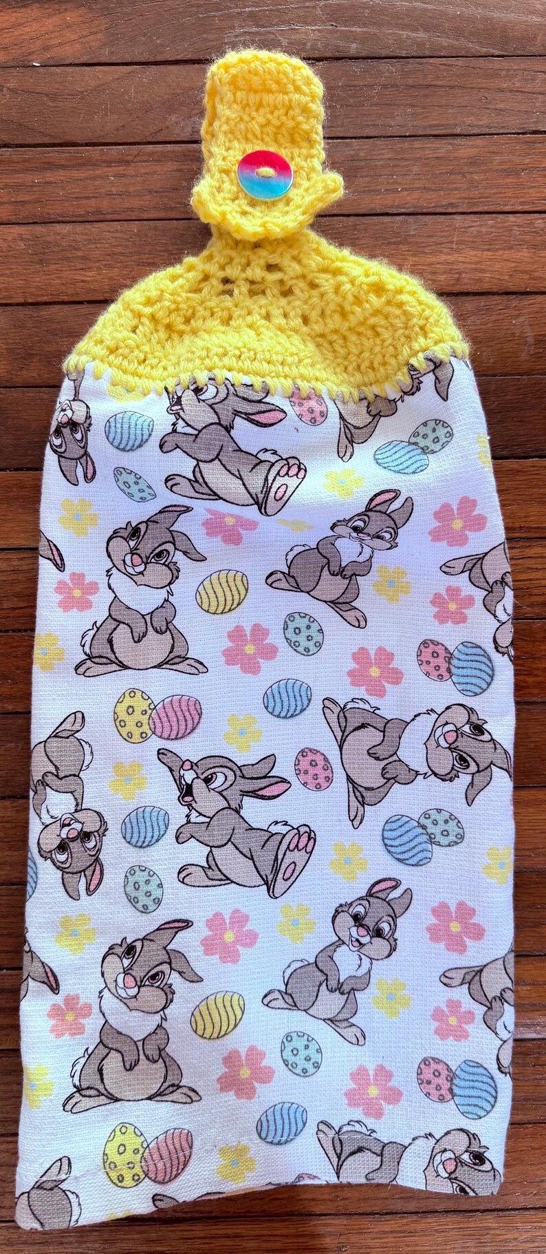 Puede incluir: Un porta-toallas de cocina de crochet amarillo con un bot&oacute;n y una toalla de cocina blanca con un patr&oacute;n de conejos marrones y huevos de Pascua coloridos.