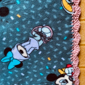 K&ouml;nnte beinhalten: Eine blaue Fleece-Decke mit einem rosa H&auml;kelrand. Die Decke zeigt ein sich wiederholendes Muster aus Konfetti und Comicfiguren, darunter Mickey Mouse und Minnie Mouse.