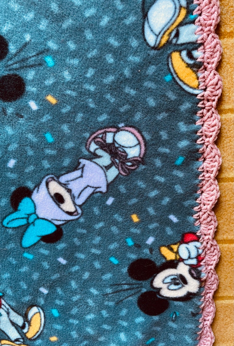 K&ouml;nnte beinhalten: Eine blaue Fleece-Decke mit einem rosa H&auml;kelrand. Die Decke zeigt ein sich wiederholendes Muster aus Konfetti und Comicfiguren, darunter Mickey Mouse und Minnie Mouse.