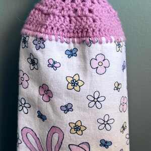 Puede incluir: Un porta-toallas de crochet morado con un bot&oacute;n azul de lunares. La toalla tiene un fondo blanco con un patr&oacute;n floral y una imagen de dibujos animados de Stitch y Angel de Lilo & Stitch.