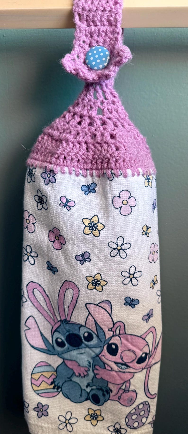 Puede incluir: Un porta-toallas de crochet morado con un bot&oacute;n azul de lunares. La toalla tiene un fondo blanco con un patr&oacute;n floral y una imagen de dibujos animados de Stitch y Angel de Lilo & Stitch.
