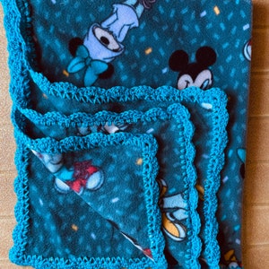 K&ouml;nnte beinhalten: Eine blaue Fleece-Decke mit Mickey-Maus-Druck und einem t&uuml;rkisfarbenen H&auml;kelrand.
