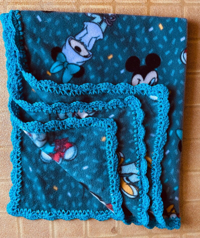 K&ouml;nnte beinhalten: Eine blaue Fleece-Decke mit Mickey-Maus-Druck und einem t&uuml;rkisfarbenen H&auml;kelrand.