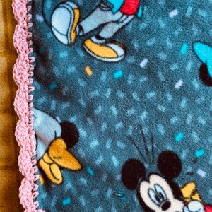 K&ouml;nnte beinhalten: Ein rosa geh&auml;kelter Rand um eine blaue Fleece-Decke mit einem Mickey-Mouse-Druck. Die Decke hat ein Konfetti-Muster und mehrere Bilder von Mickey Mouse.
