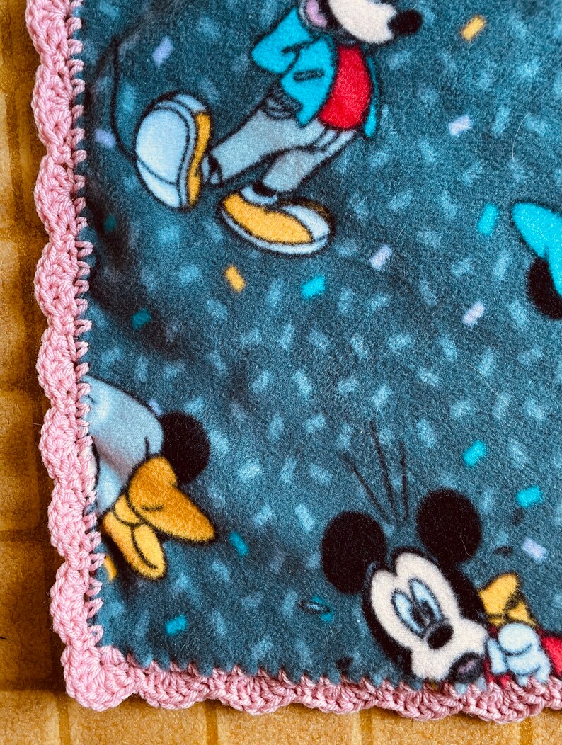 K&ouml;nnte beinhalten: Ein rosa geh&auml;kelter Rand um eine blaue Fleece-Decke mit einem Mickey-Mouse-Druck. Die Decke hat ein Konfetti-Muster und mehrere Bilder von Mickey Mouse.