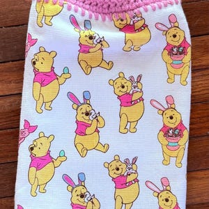 Puede incluir: Un pa&ntilde;o de cocina de crochet rosa con un lazo de bot&oacute;n y un panel de tela blanca con un patr&oacute;n repetido de personajes de Winnie the Pooh vestidos con orejas de conejo que sostienen huevos de Pascua y un conejito.