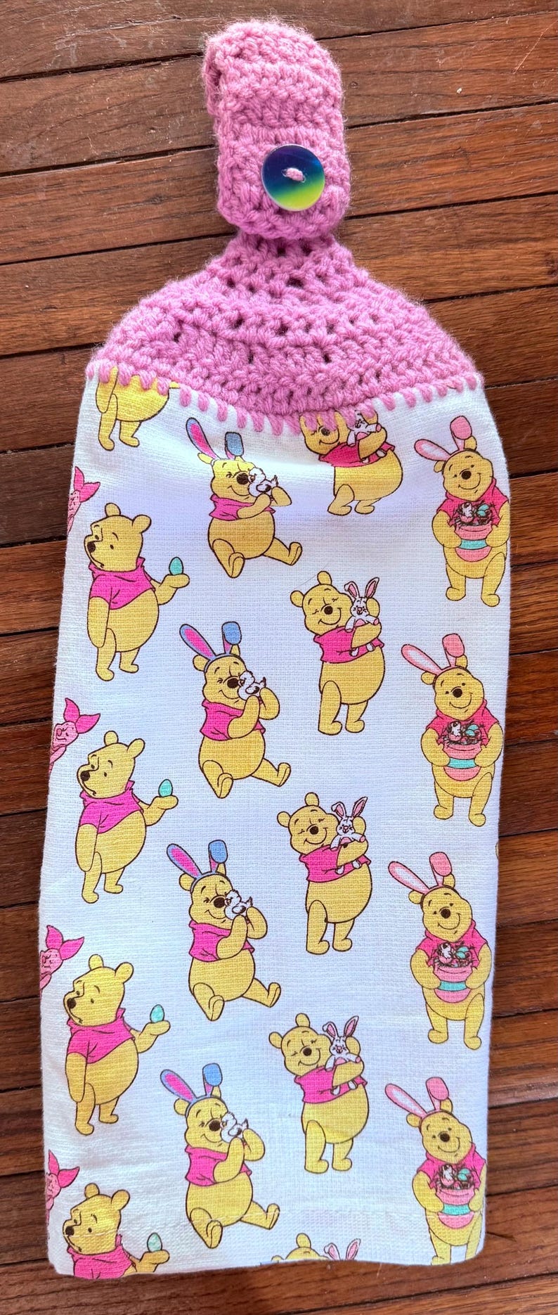 Puede incluir: Un pa&ntilde;o de cocina de crochet rosa con un lazo de bot&oacute;n y un panel de tela blanca con un patr&oacute;n repetido de personajes de Winnie the Pooh vestidos con orejas de conejo que sostienen huevos de Pascua y un conejito.