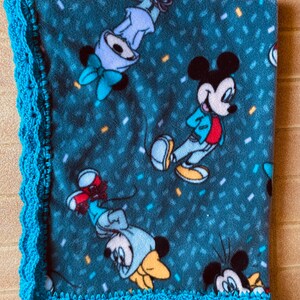 K&ouml;nnte beinhalten: Eine blaue Fleece-Decke mit einem geh&auml;kelten t&uuml;rkisfarbenen Rand. Die Decke zeigt ein sich wiederholendes Muster von Mickey Mouse und Minnie Mouse Figuren.