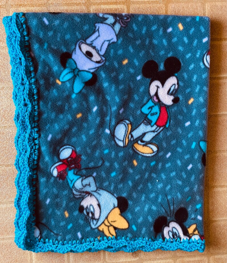 K&ouml;nnte beinhalten: Eine blaue Fleece-Decke mit einem geh&auml;kelten t&uuml;rkisfarbenen Rand. Die Decke zeigt ein sich wiederholendes Muster von Mickey Mouse und Minnie Mouse Figuren.