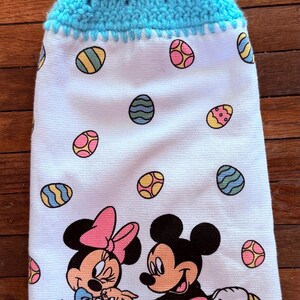 Puede incluir: Una toalla de cocina blanca con un lazo de colgar de crochet azul y un bot&oacute;n. La toalla presenta un patr&oacute;n de huevos de Pascua coloridos y una imagen de dibujos animados de Mickey y Minnie Mouse sosteniendo cestas de Pascua.
