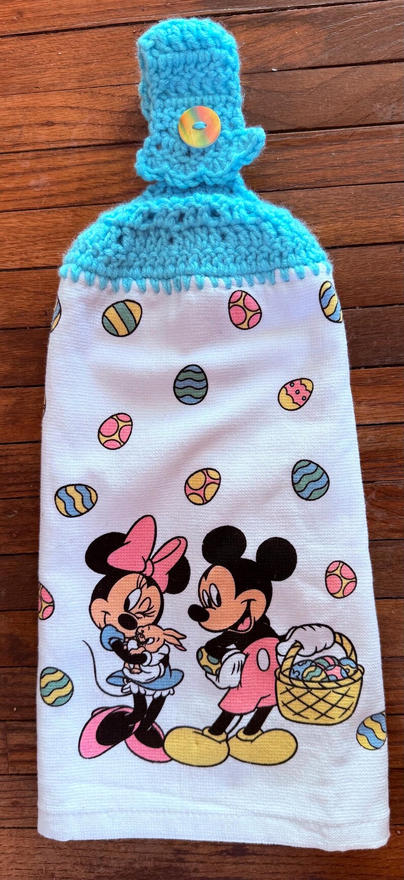 Puede incluir: Una toalla de cocina blanca con un lazo de colgar de crochet azul y un bot&oacute;n. La toalla presenta un patr&oacute;n de huevos de Pascua coloridos y una imagen de dibujos animados de Mickey y Minnie Mouse sosteniendo cestas de Pascua.