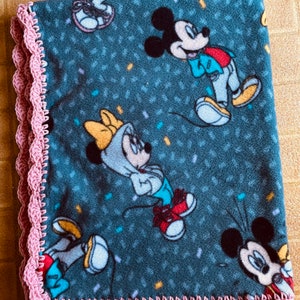 K&ouml;nnte beinhalten: Eine weiche, blaue Fleece-Decke mit einem rosa H&auml;kelrand. Die Decke zeigt ein sich wiederholendes Muster von Mickey und Minnie Mouse in verschiedenen Posen.