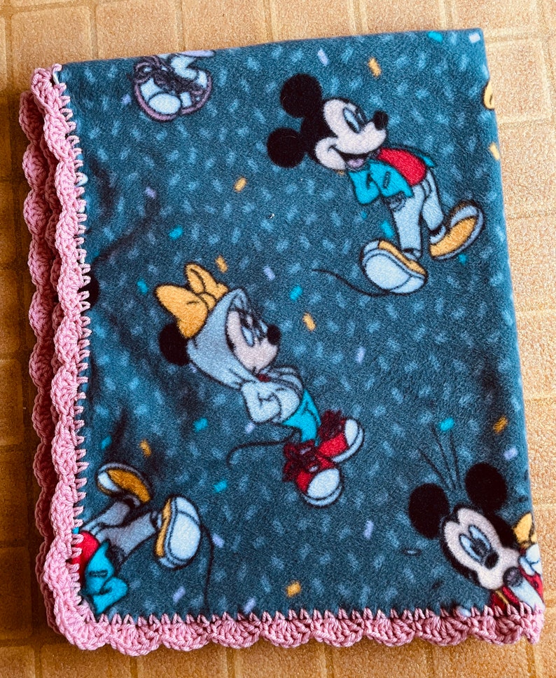 K&ouml;nnte beinhalten: Eine weiche, blaue Fleece-Decke mit einem rosa H&auml;kelrand. Die Decke zeigt ein sich wiederholendes Muster von Mickey und Minnie Mouse in verschiedenen Posen.