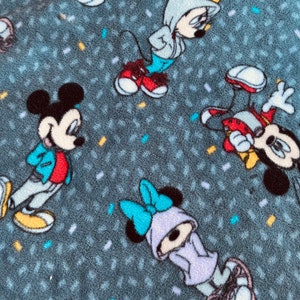 K&ouml;nnte beinhalten: Eine graue Fleece-Decke mit einem sich wiederholenden Muster von Mickey Mouse und Minnie Mouse Figuren, die verschiedene farbige Outfits tragen. Die Figuren tragen Turnschuhe und um sie herum sind Konfetti verstreut.