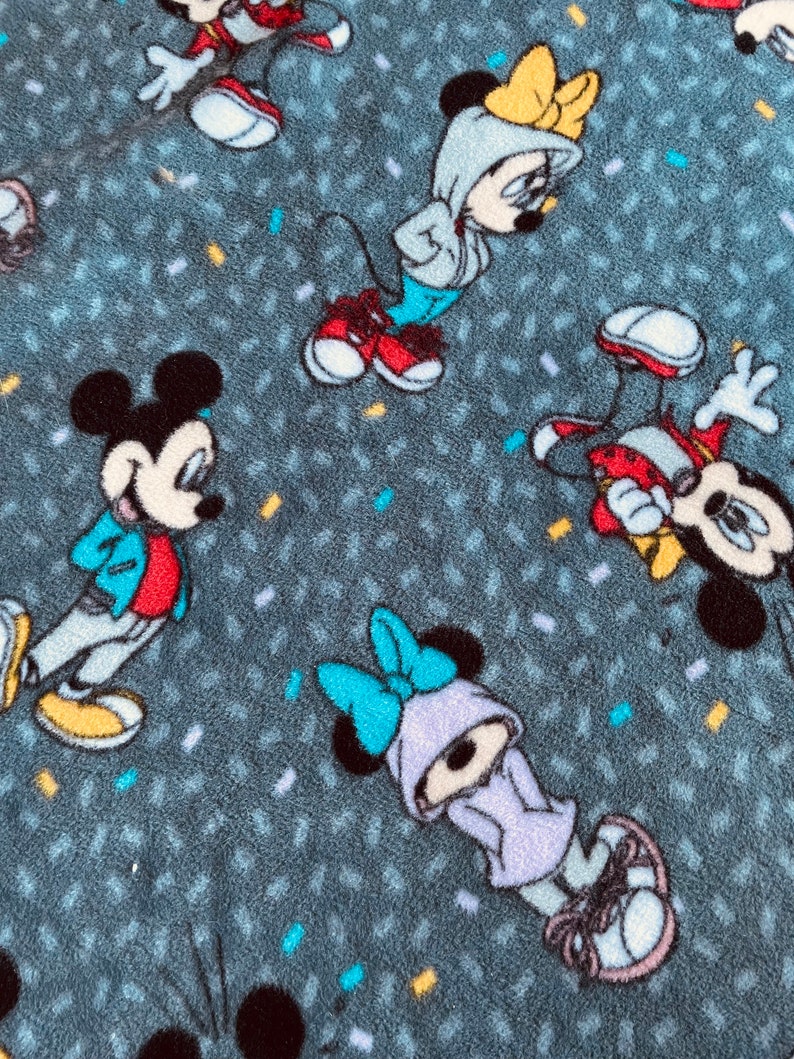 K&ouml;nnte beinhalten: Eine graue Fleece-Decke mit einem sich wiederholenden Muster von Mickey Mouse und Minnie Mouse Figuren, die verschiedene farbige Outfits tragen. Die Figuren tragen Turnschuhe und um sie herum sind Konfetti verstreut.