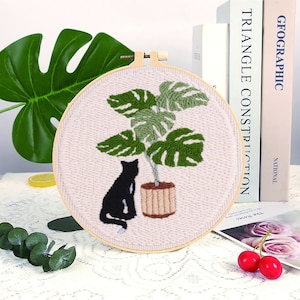 Op de afbeelding: Een zwarte kat die naast een groene plant in een pot met grote bladeren zit. De afbeelding is geborduurd op een witte stof die over een houten borduurring is gespannen.