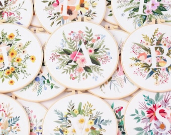 May include: A collection of 10 embroidery hoops with floral designs and a letter in the center of each hoop. The letters are A, B, E, F, P, S, V, and W. The hoops are all white with a wooden frame.