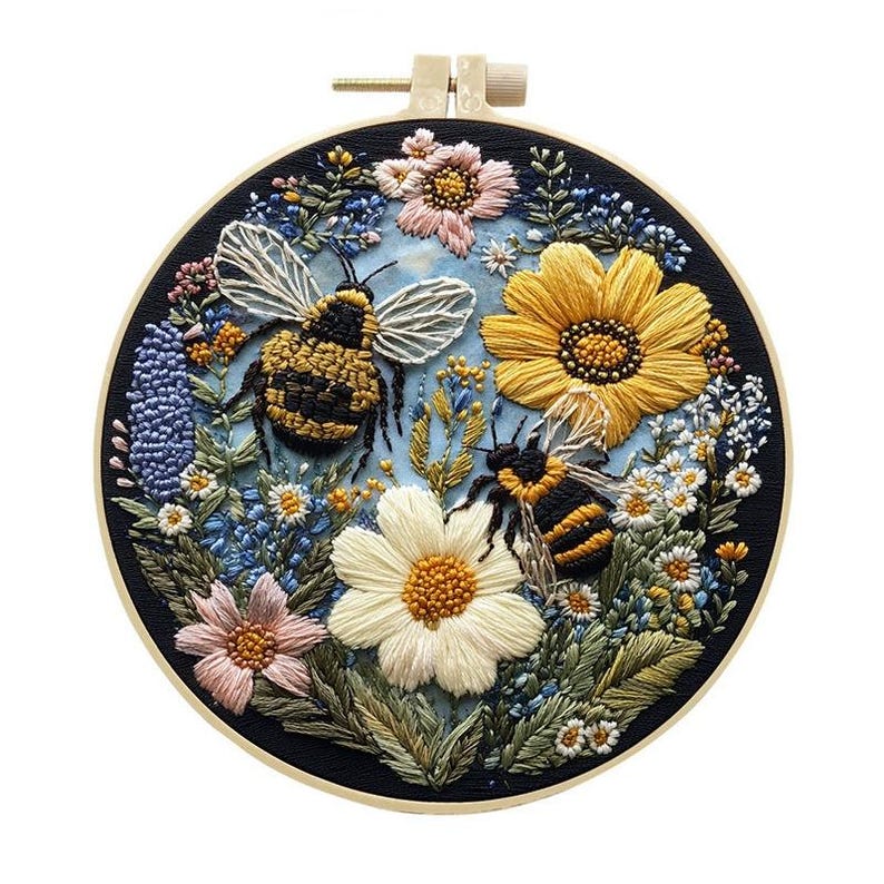May include: Embroidered hoop art featuring a detailed floral and bee design. The artwork showcases a black background with colorful flowers, including yellow sunflowers, pink and white daisies, and blue wildflowers. Two bees are prominently displayed.