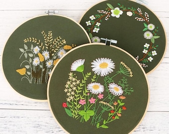 May include: Three embroidery hoops with green fabric and floral embroidery designs. The designs include a wreath of white flowers, a cluster of white daisies, and a bouquet of white daisies with pink and red accents.