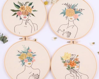 Könnte beinhalten: Vier Stickrahmen mit beigefarbenem Stoff und Holzrahmen. Jeder Rahmen zeigt eine Linienzeichnung einer Person mit Blumen, die ihr Gesicht bedecken. Die Blumen sind in Gelb-, Orange-, Rosa- und Grüntönen gestickt.