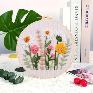 May include: A circular embroidery hoop with a floral design. The embroidery depicts flowers in white, pink, and yellow, with green leaves and stems. The hoop is made of light-coloured wood, and the fabric is a light beige.