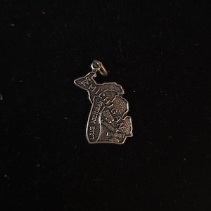 Puede incluir: Un dije de metal plateado con la forma del estado de Michigan. La superficie presenta texto en relieve que dice MICHIGAN, LAKE MICHIGAN, GRAND RAPIDS, LANSING y DETROIT, junto con pequeños iconos de un ciervo, una estrella y un automóvil.