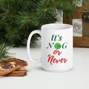 May include: White ceramic mug with the text "It's NOG or Never" in green and red. A green ornament graphic is between the words "NOG". The mug has a curved handle and is set against a background of Christmas greenery.