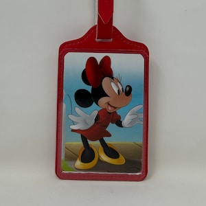 May include: Red leather luggage tag with a picture of Minnie Mouse in a red dress and yellow shoes. The tag has a silver buckle.