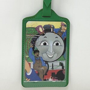 May include: A green luggage tag featuring a rectangular frame with a silver buckle strap. The center displays a colorful illustration of a smiling grey train engine face surrounded by human figures in blue and brown overalls against a green landscape.