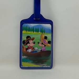 May include: Blue luggage tag featuring a picture of Mickey Mouse and Minnie Mouse in a boat on a lake. The tag has a blue strap with a silver buckle.