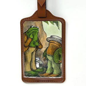 May include: A rectangular luggage tag with a brown leather-like border and a matching adjustable strap featuring a silver-tone metal buckle. The center displays a printed illustration of two anthropomorphic frogs wearing clothing.