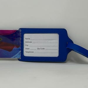May include: Blue luggage tag with a clear plastic insert featuring a cartoon image of a girl with long blonde hair wearing a pink and blue dress. The tag has a white label with the words "Name", "Address", "City", "State", "Zip Code", and "Telephone".