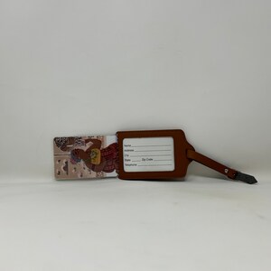 Pode incluir: Uma etiqueta de bagagem de couro castanho com uma correia e fivela. A etiqueta tem uma janela retangular para informa&ccedil;&otilde;es de nome e endere&ccedil;o. Uma inser&ccedil;&atilde;o decorativa apresenta uma ilustra&ccedil;&atilde;o colorida de uma pessoa.