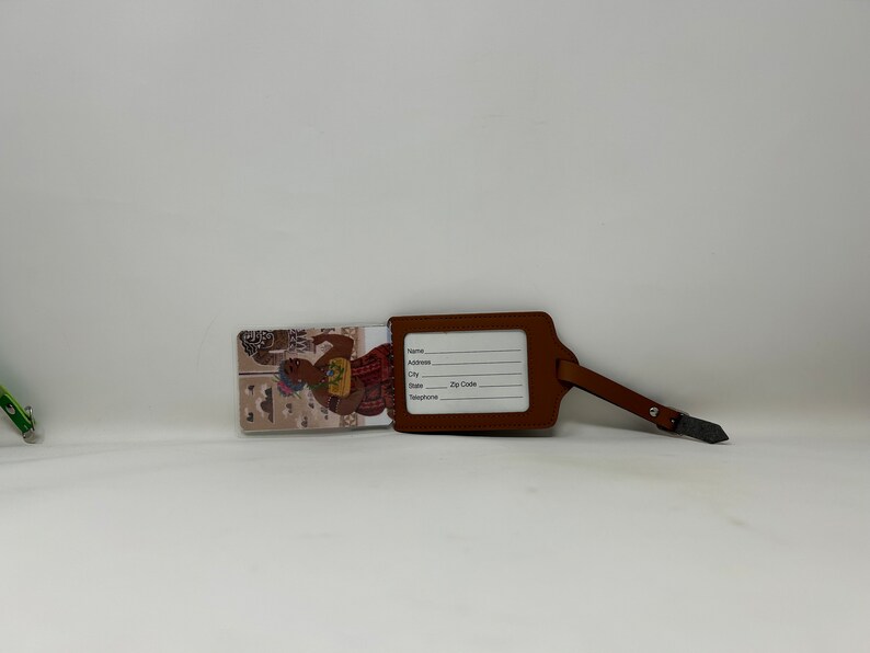 Pode incluir: Uma etiqueta de bagagem de couro castanho com uma correia e fivela. A etiqueta tem uma janela retangular para informa&ccedil;&otilde;es de nome e endere&ccedil;o. Uma inser&ccedil;&atilde;o decorativa apresenta uma ilustra&ccedil;&atilde;o colorida de uma pessoa.