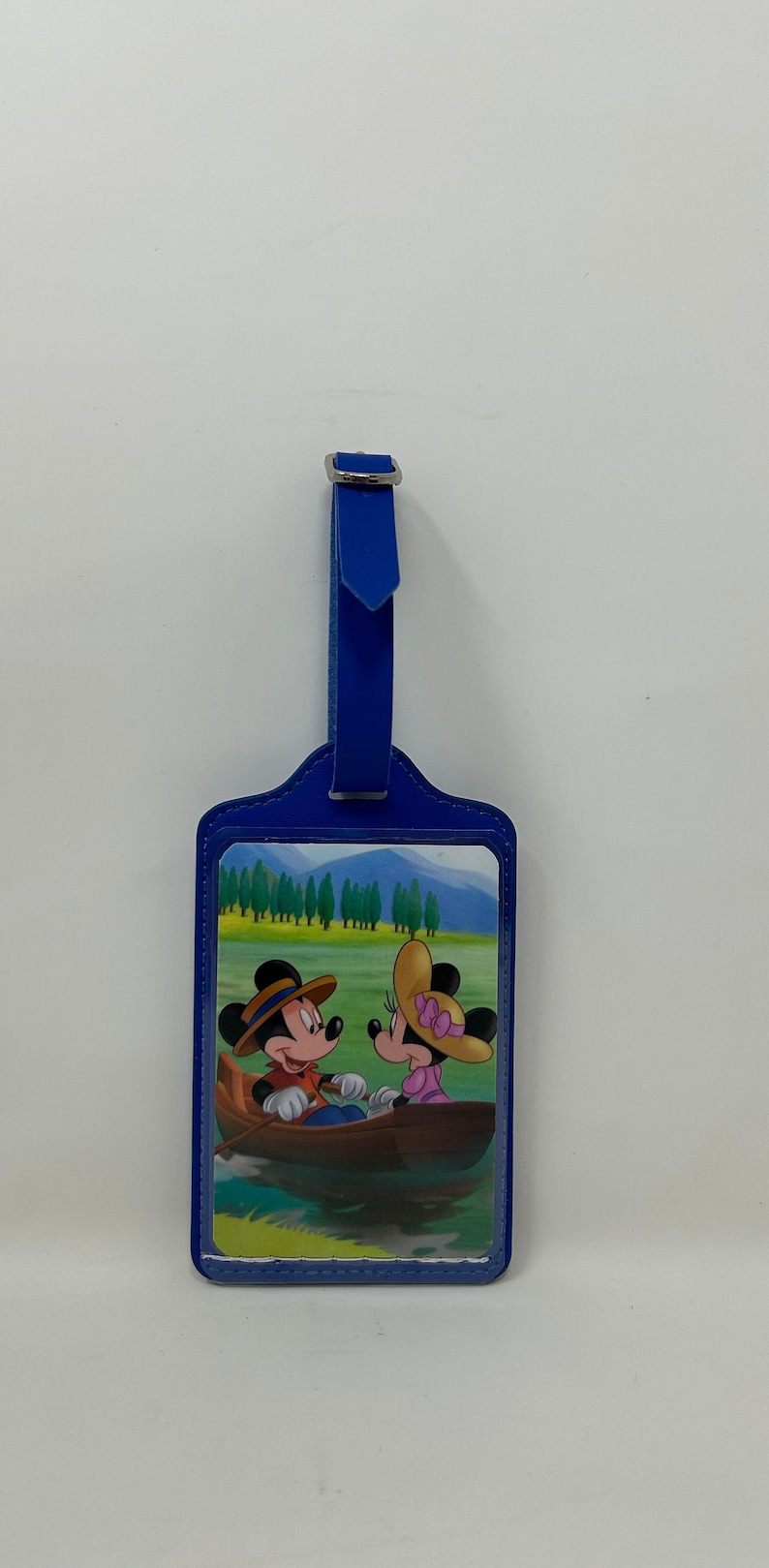 May include: Blue luggage tag featuring a picture of Mickey Mouse and Minnie Mouse in a boat on a lake. The tag has a blue strap with a silver buckle.