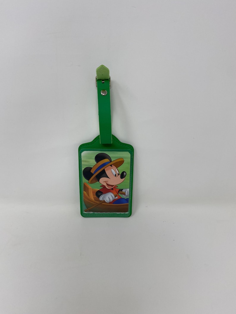 Peut inclure: Etiquette &agrave; bagages verte avec une image de Mickey Mouse portant un chapeau et ramant dans un bateau. L'&eacute;tiquette a une sangle verte.