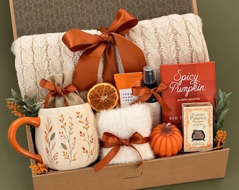 May include: A gift box containing a cream-coloured cable knit blanket with a rust-coloured ribbon, a pumpkin-shaped mug with leaf patterns, a small burlap bag, a dried orange slice, hand cream, a spray bottle, Spicy Pumpkin tea, and pumpkin soap.