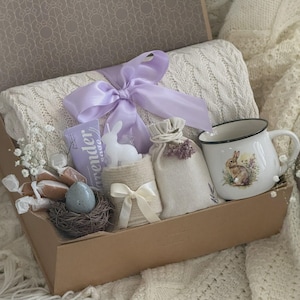 May include: A gift box filled with a white blanket, a lavender-scented soap, a bunny-shaped candle, a mug with a bunny illustration, a small bag, and a decorative nest with an egg. The box is tied with a lavender ribbon.