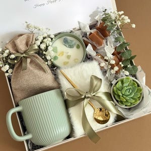 May include: A white gift box filled with a green mug, a small potted succulent, a white fuzzy blanket, a gold spoon, a small bag, a candle with crystals, and caramel candies. The box is decorated with white flowers and green leaves. The text "The Botanical Box" is printed on the bottom of the box.