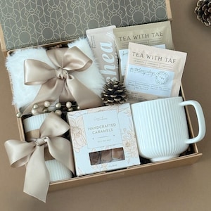 May include: A gift box containing a white faux fur item tied with a champagne ribbon, a white ribbed ceramic mug, a box of handcrafted caramels with sea salt, and two Tea With Tae tea sachets. The box interior features a geometric pattern.