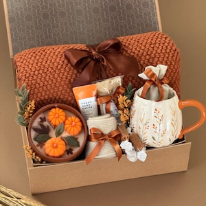 May include: A gift box containing a rust-colored knit blanket with a brown ribbon, a ceramic mug with a leaf pattern and orange handle, a candle with three pumpkin-shaped wax inserts, a tube of vegan hand cream, a small beige pouch, and socks.