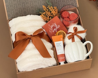 May include: A gift box containing a white plush blanket tied with a copper ribbon, a Spicy Pumpkin candle, a tube of Voesh Vegan Hand Creme, a white pumpkin-shaped ceramic mug, dried orange slices, and yellow floral accents.