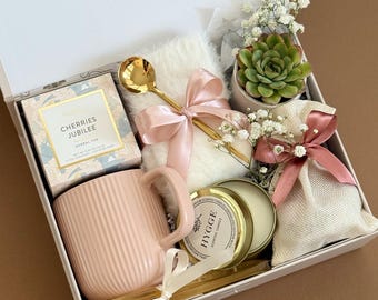 May include: A gift box filled with pink items, including a mug, candle, tea, and a succulent plant. The box is decorated with a pink ribbon and a gold spoon. The text "A Touch of Pink" is visible.