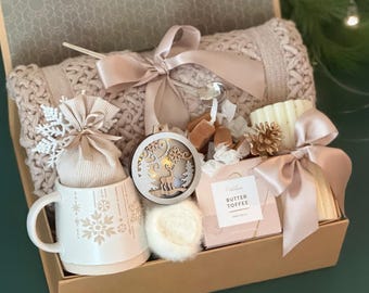 May include: A gift box filled with a beige knitted blanket, a white mug with snowflake designs, a decorative ornament, a candle, and a box of toffee. The items are tied with satin ribbons and bows, creating a cozy and festive presentation.